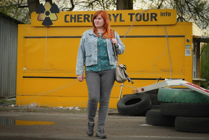 身穿牛仔夹克的红发女子站在一面黄色横幅旁，看上去若有所思 "切尔诺贝利之旅"