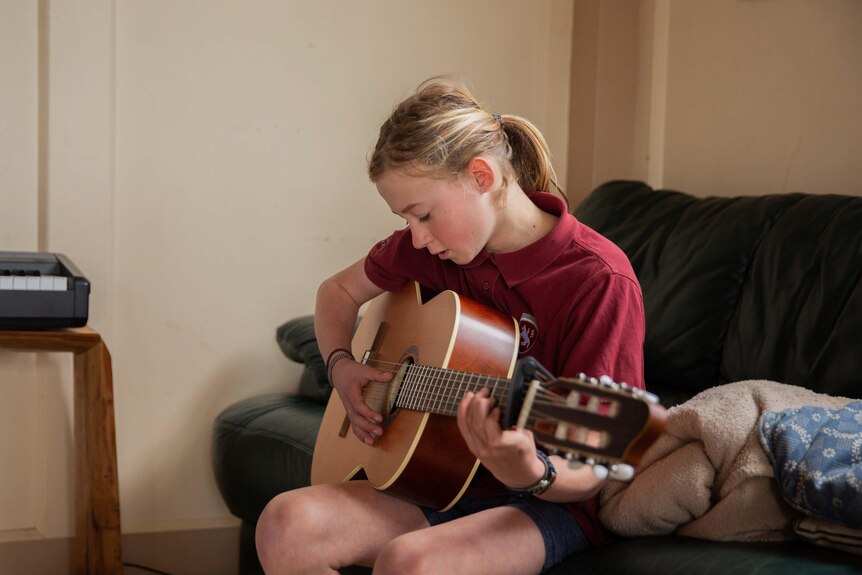 A boy in a maroon polo top sitting on a couch playing guitar.