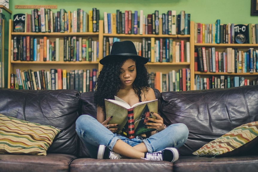 Girl sitting on couch reading a book with book shelves in the background.