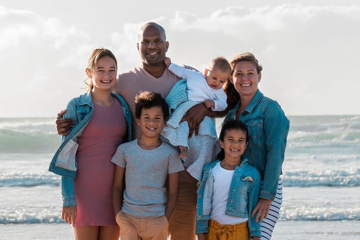 A young family of five stands in the sun in front of a beach.