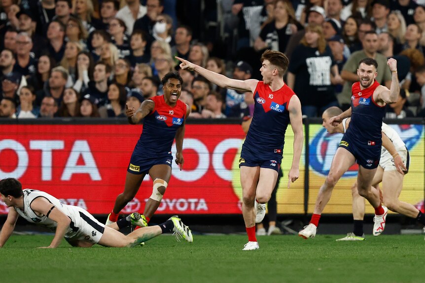 A Melbourne AFL player points in celebration after a goal as his grinning teammates run towards him.