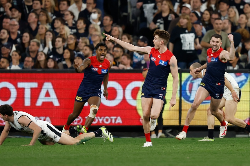 A Melbourne AFL player points in celebration after a goal as his grinning teammates run towards him.