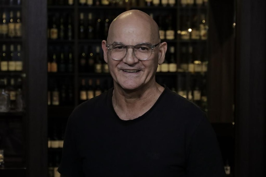 Bald man with glasses smiles at camera in front of wine bottles