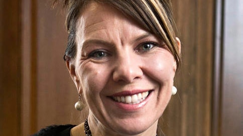 A woman smiles while looking directly at the camera with her hair pinned back, wearing pearl earrings and a necklace