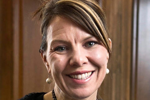 A woman smiles while looking directly at the camera with her hair pinned back, wearing pearl earrings and a necklace