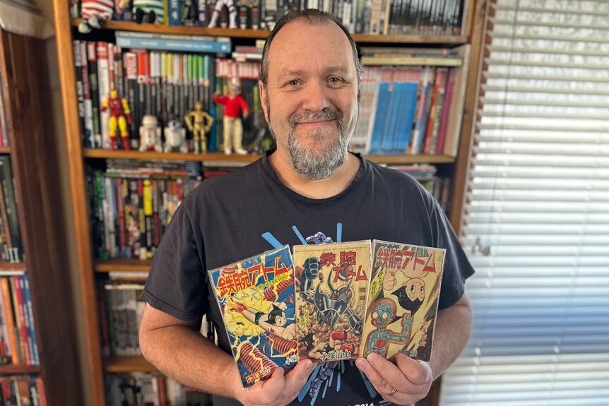 A man with a pony tail and beard holds three small, rare comics in his hands, standing in front of a bookshelf full of comics