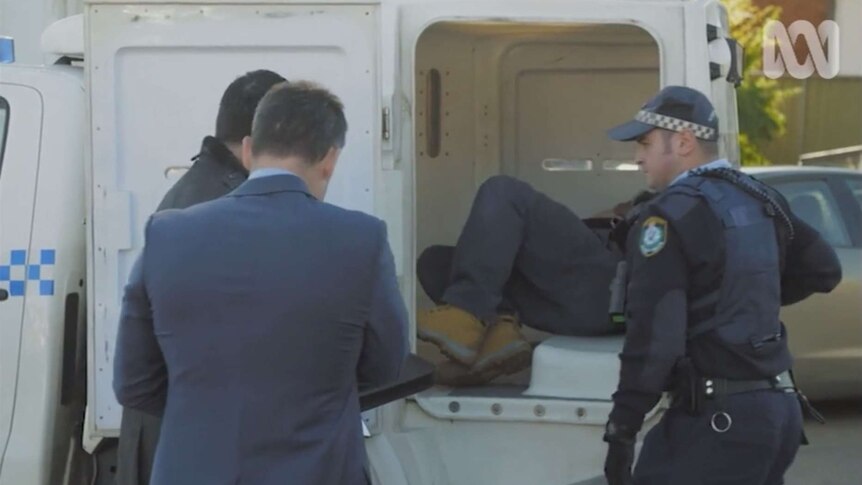 A man lays in a police van, as several other people stand nearby.