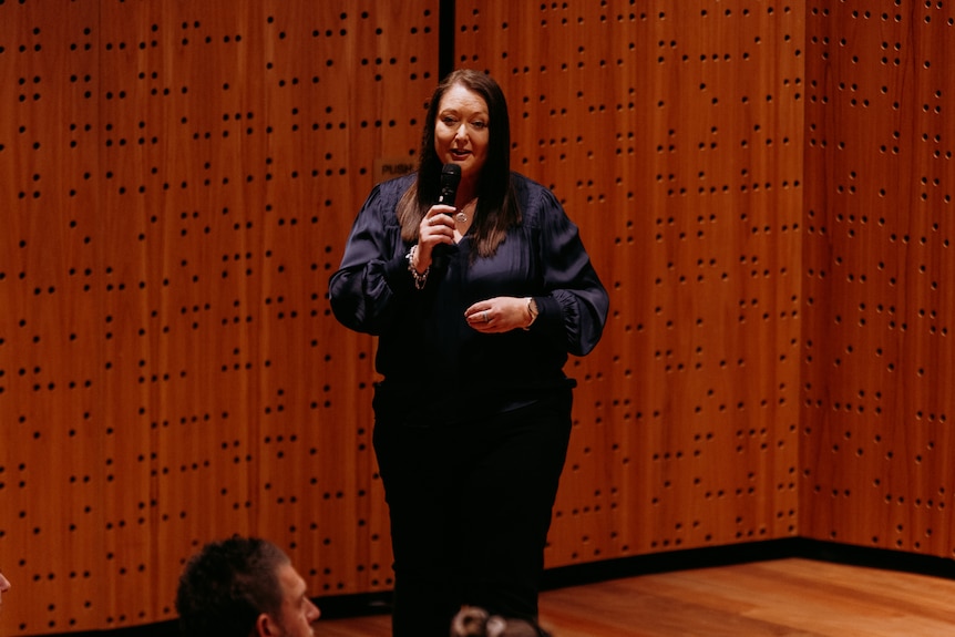 Lisa Parrello, a middle-aged white woman with dark brown hair, wears a navy blouse and stands on a stage speaking into a mic.