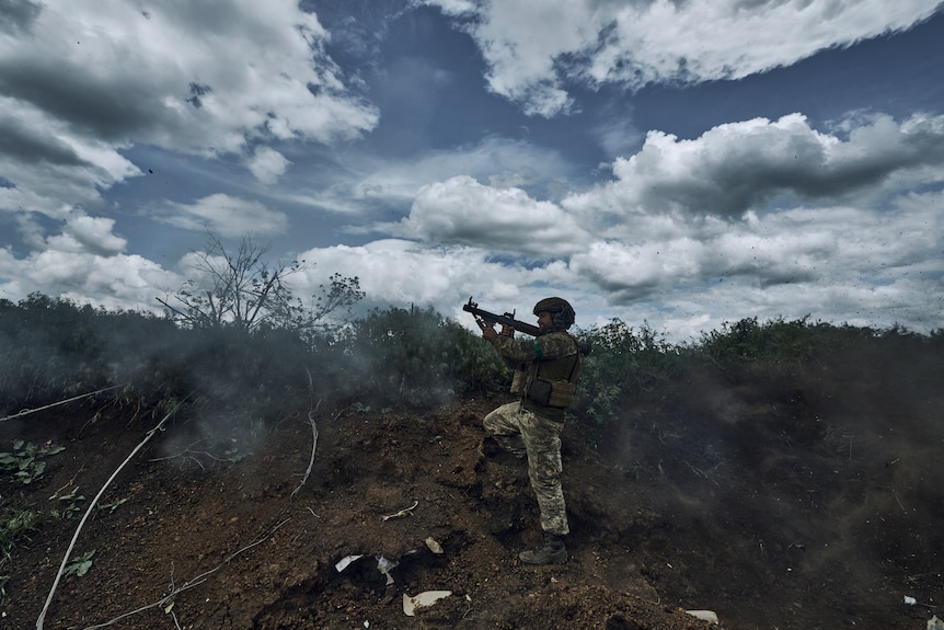 A soldier stands on a small rise of ground and fires a shoulder mounted, cylindrical rpg.