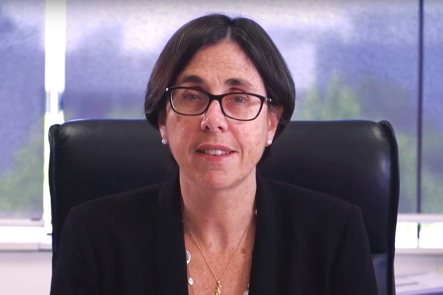 A woman with a short haircut and black glasses sits at a desk looking directly at camera