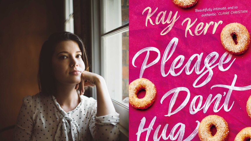 Kay Kerr, author of Please Don't Hug Me, stands next to the cover of her book. It is pink with cinnamon donuts on it.