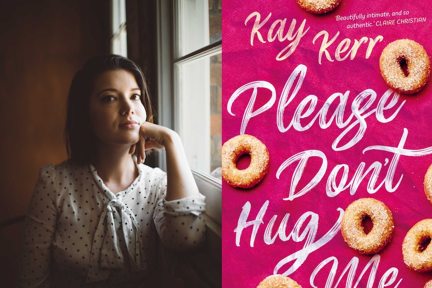 Kay Kerr, author of Please Don't Hug Me, stands next to the cover of her book. It is pink with cinnamon donuts on it.