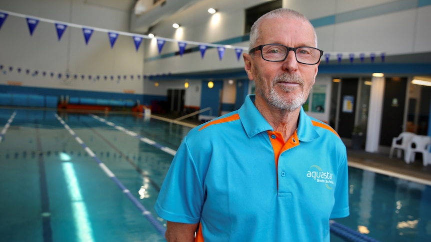 Steve is standing in front of an indoor laned swimming pool wearing a blue "Aquastart Swim School" shirt and smiling.
