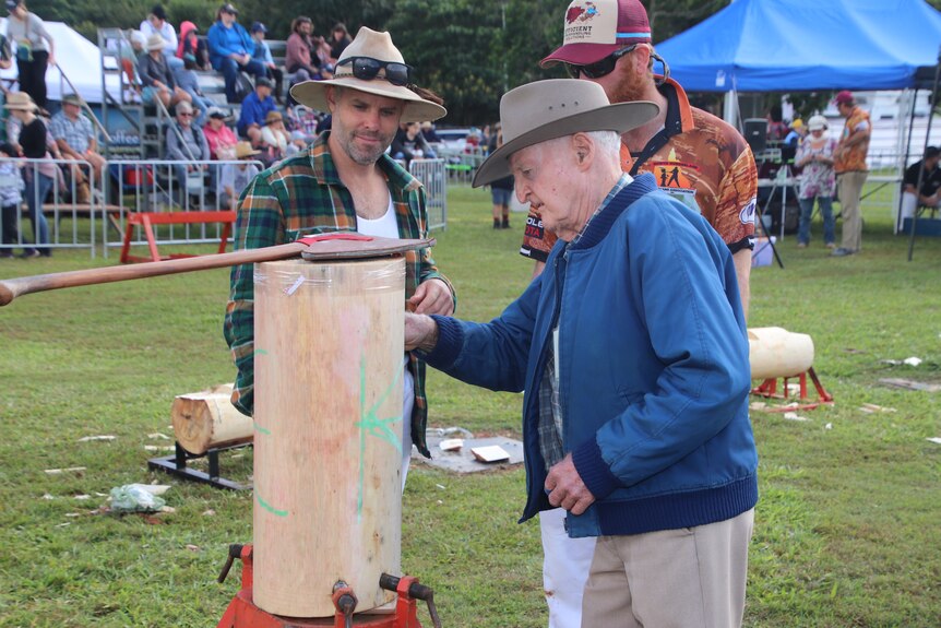 An old man points to a piece of wood with an axe on top as two others look on.