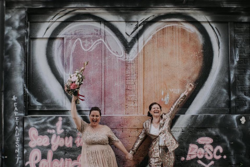 Ms Mackie and Ms Clark hold hands in their wedding outfits in front of a graffitied heart.