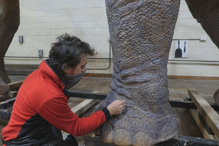 A man is kneeling painting the foot of a large blue dinosaur