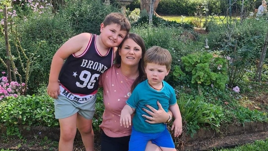 A woman smiles in her back yard with her two sons, one school aged and one a toddler