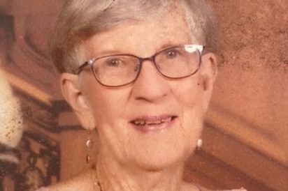 A elderly woman with short grey hair and small round glasses, wearing a pink top, smiling.
