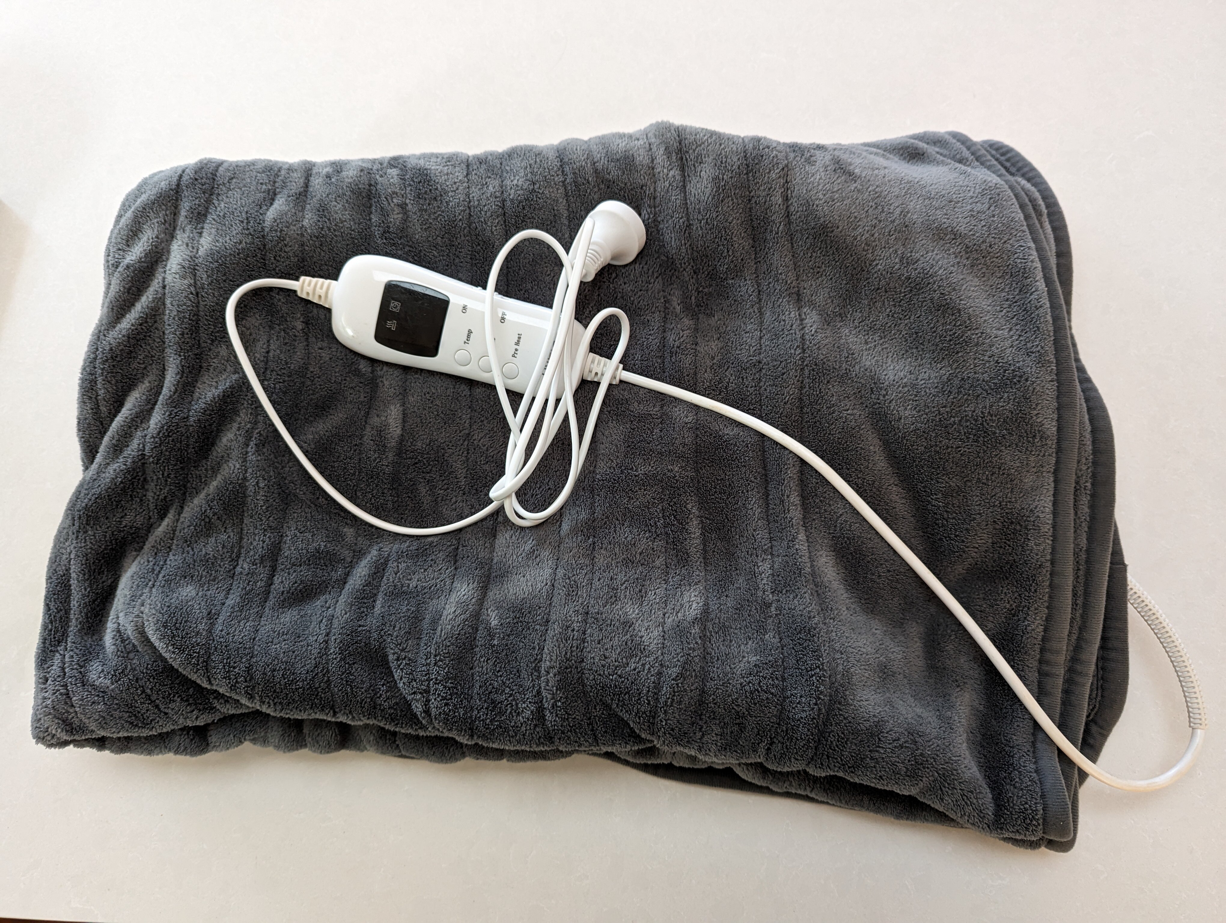 A fluffy grey heated throw blanket is folded on a white surface, with the power cord gathered on top.