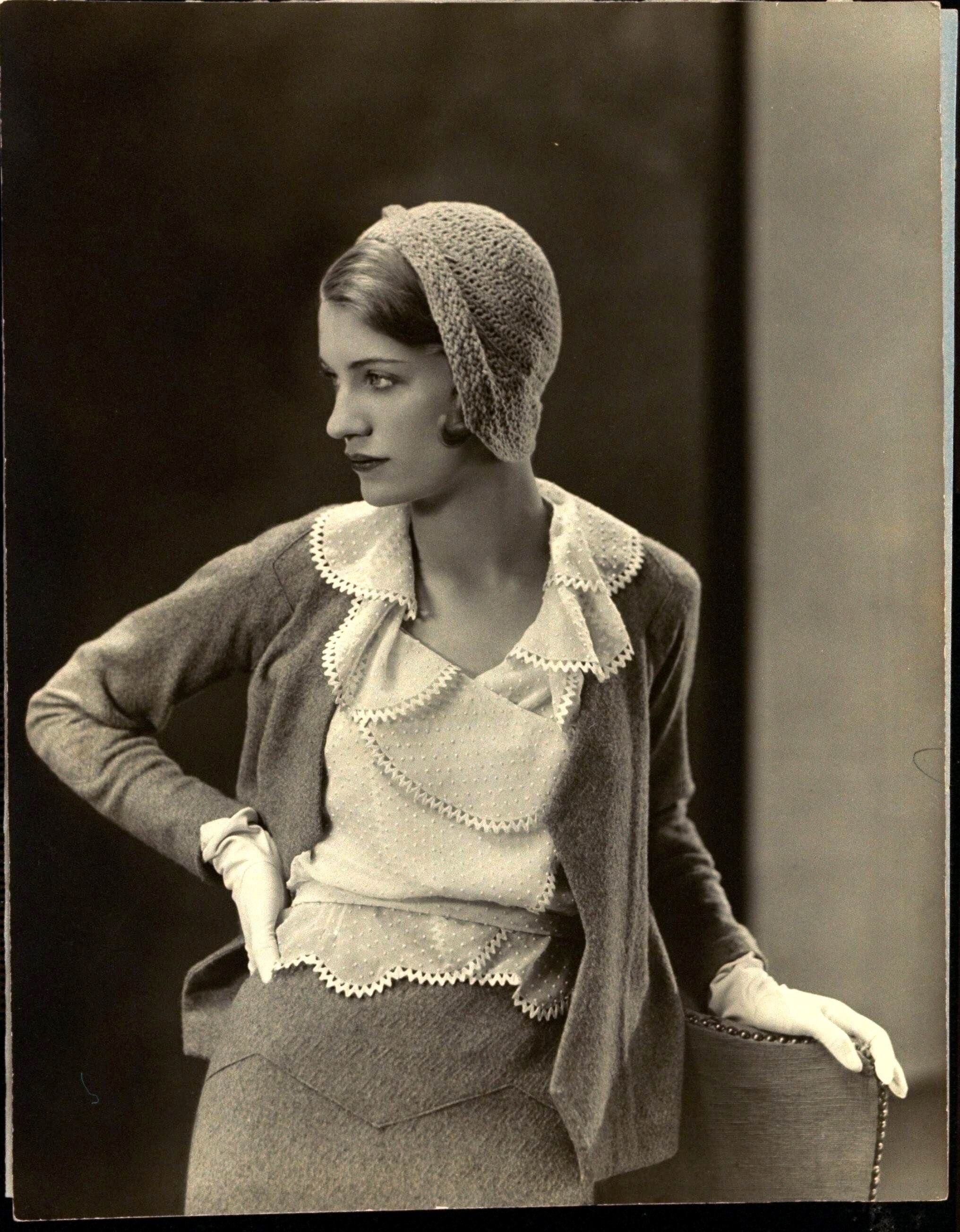 A black and white photo of a young woman wearing 1930s attire looking to the side, with her gloved hand on a chair