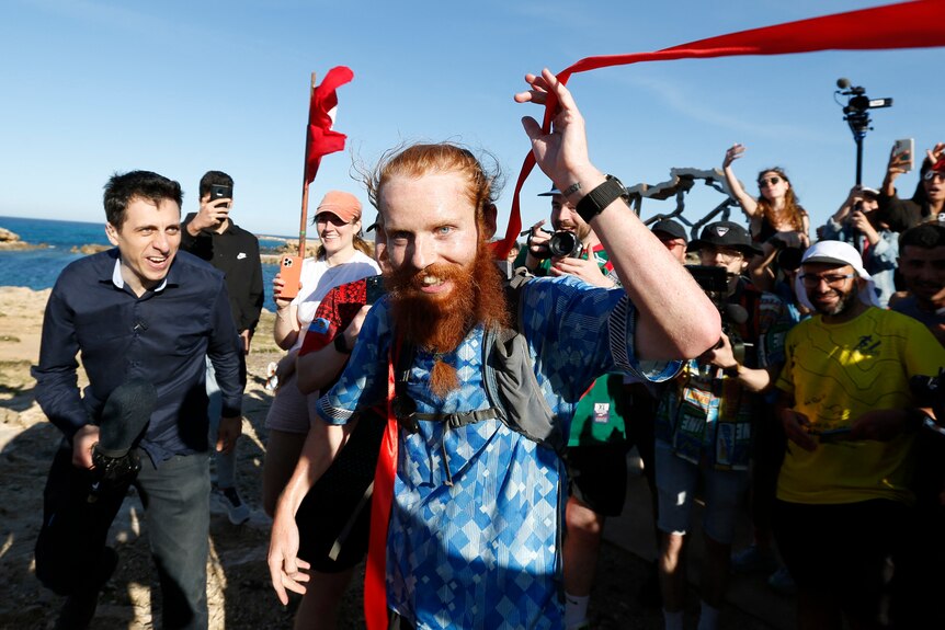 A red haired man in a blue shirt corssing a finishing ribbon surrounded by a crowd. 