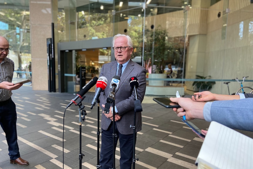 A man in glasses and a suit stands behind a row of microphones