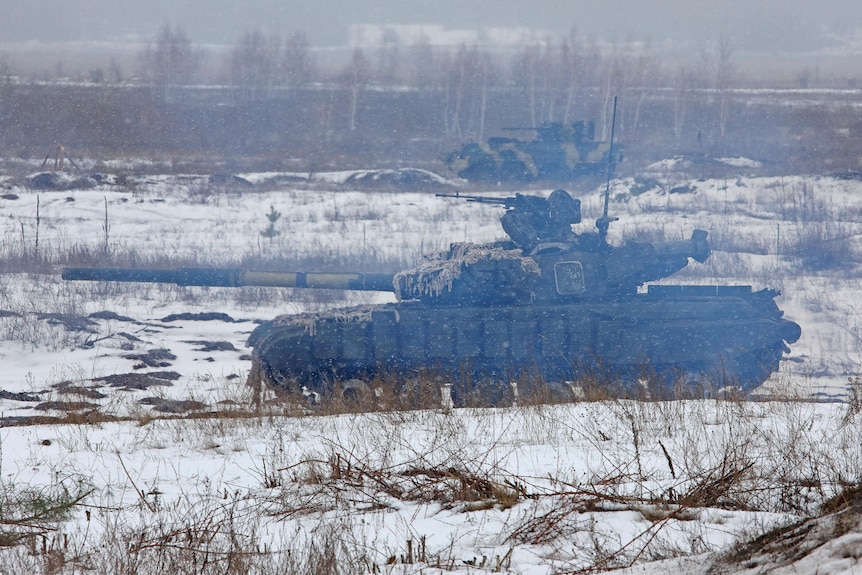 A grey military tank drives through the snow on a wintry field on a hazy day.