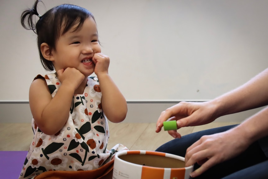 A young girl smiles off camera as she plays with blocks. Her hair is in pigtails.