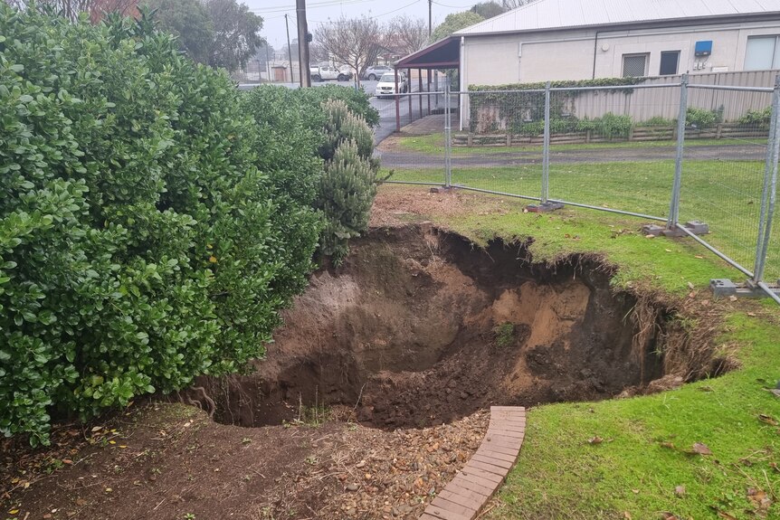 A sinkhole surrounded by temporary fencing, with a hedge to the left of the hole.
