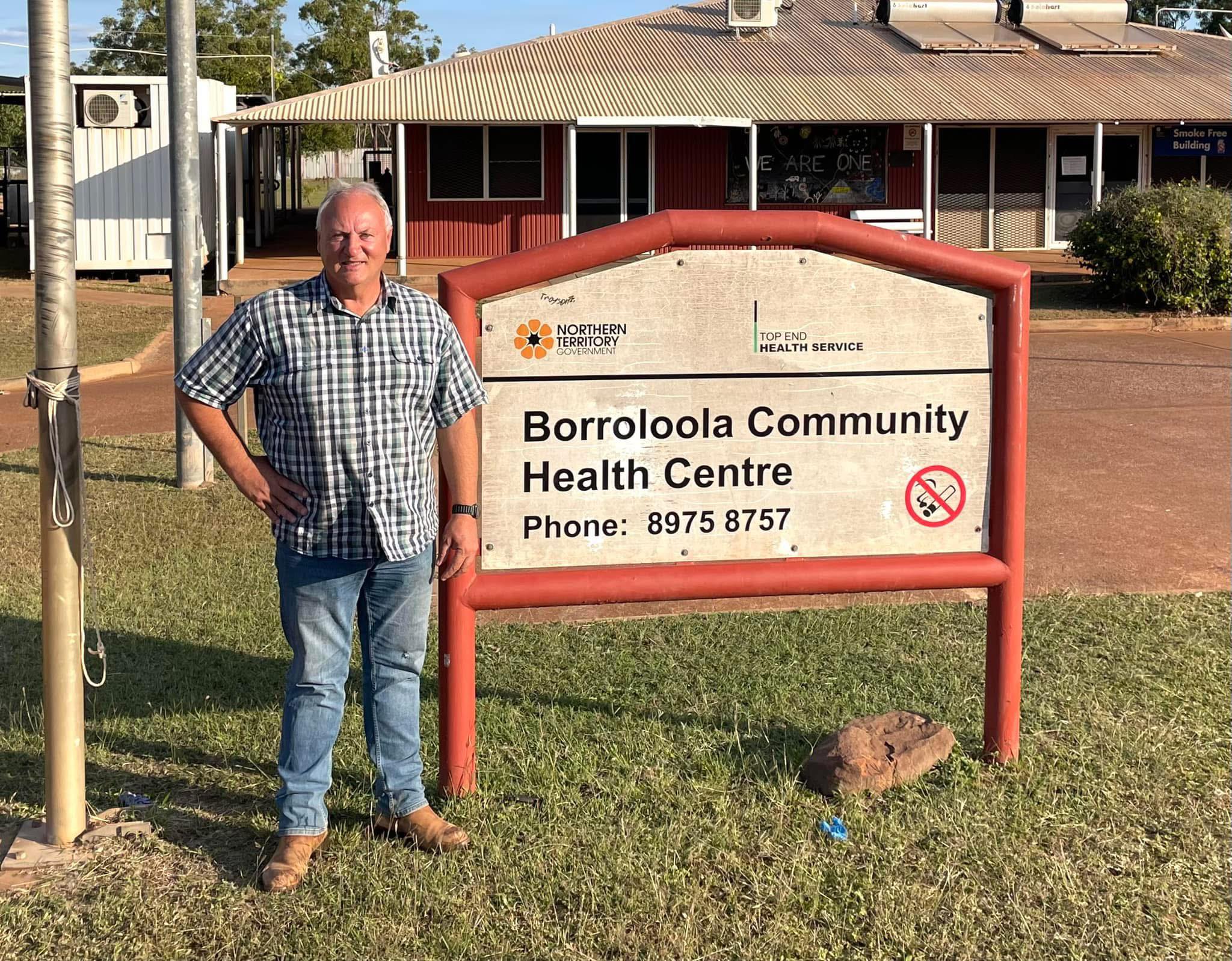 White man in plaid shirt and jeans standing in front of sign which reads 'Borroloola Community Health Clinic'.