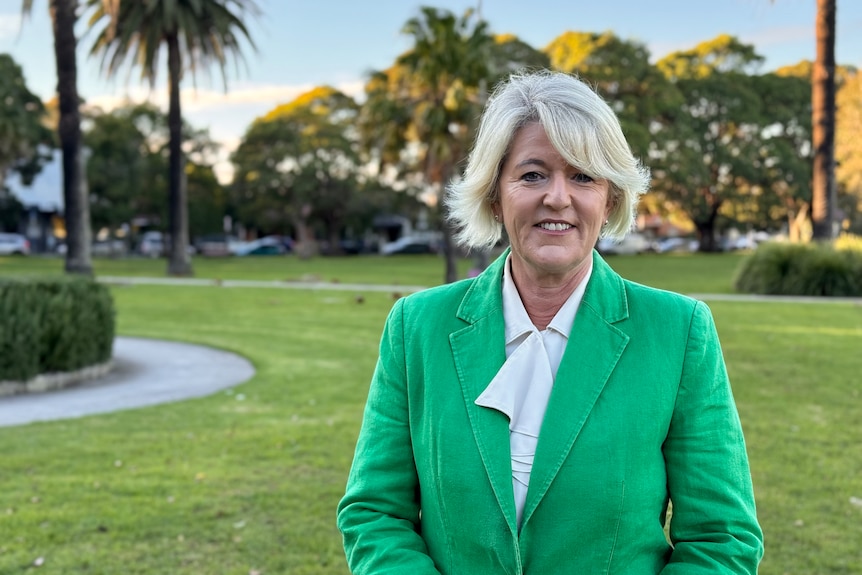 A middle-aged woman in a bright green blazer stands in a park and smiles at the camera during the early morning.