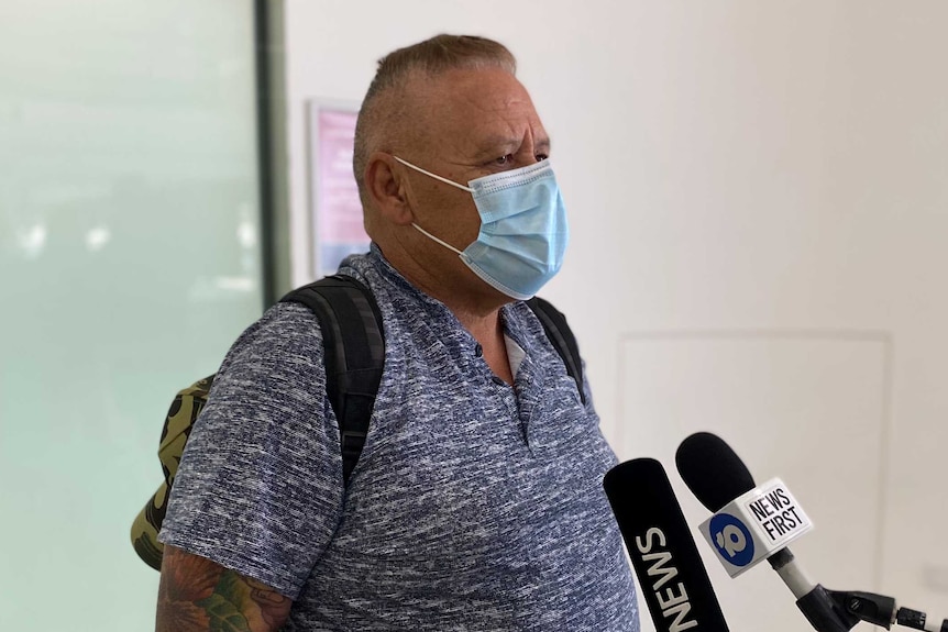 A man wearing a mask and a blue shirt talking to media after arriving at Perth airport from Sydney