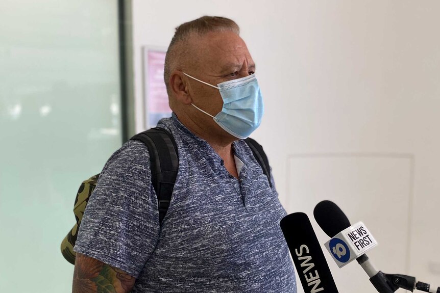 A man wearing a mask and a blue shirt talking to media after arriving at Perth airport from Sydney