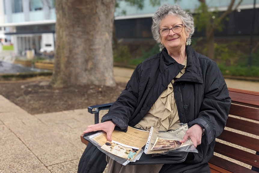 A smiling woman sitting on a park bench and holding a folder of photos and old paperwork and newspaper clippings 