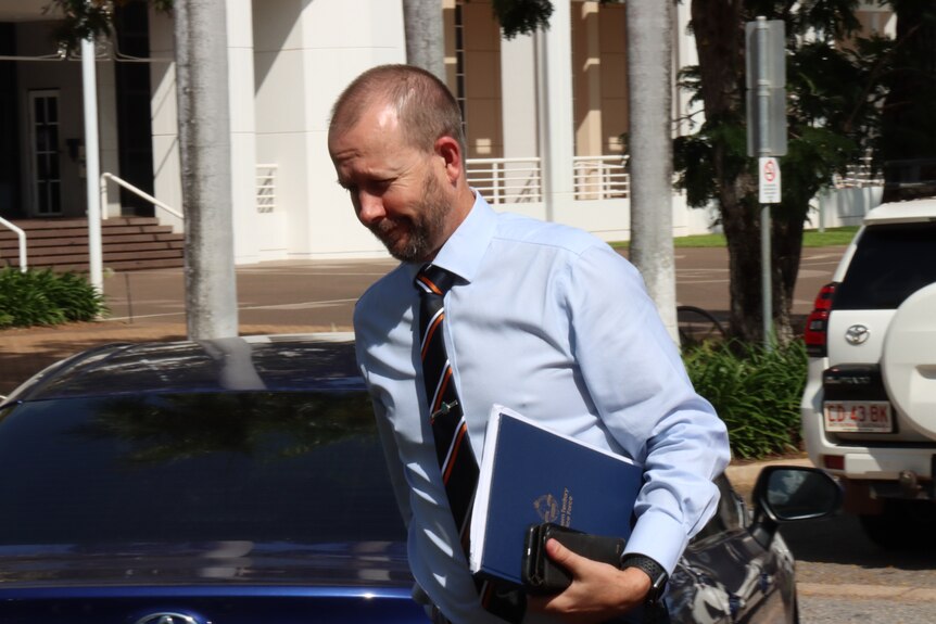 A man in a blue shirt and tie and holding files under his arm walking, in front of the NT Supreme Court building. 