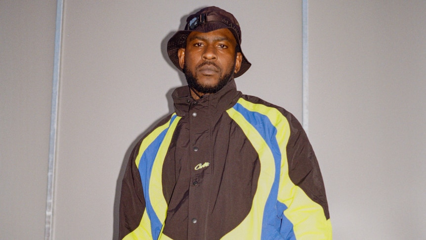 Skepta stands against an off-white wall in a brown bucket hat, brown, blue and yellow jacket and camoflage pants