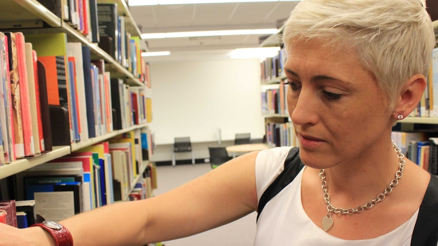 A woman with short blond hair, white t-shirt looks at books in a library.