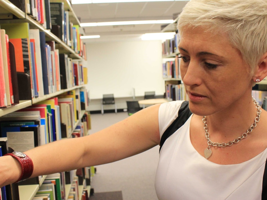 A woman with short blond hair, white t-shirt looks at books in a library.