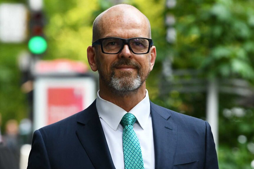 Simon Overland, wearing glasses, a dark suit and green tie, smiles as he stands on a city street.