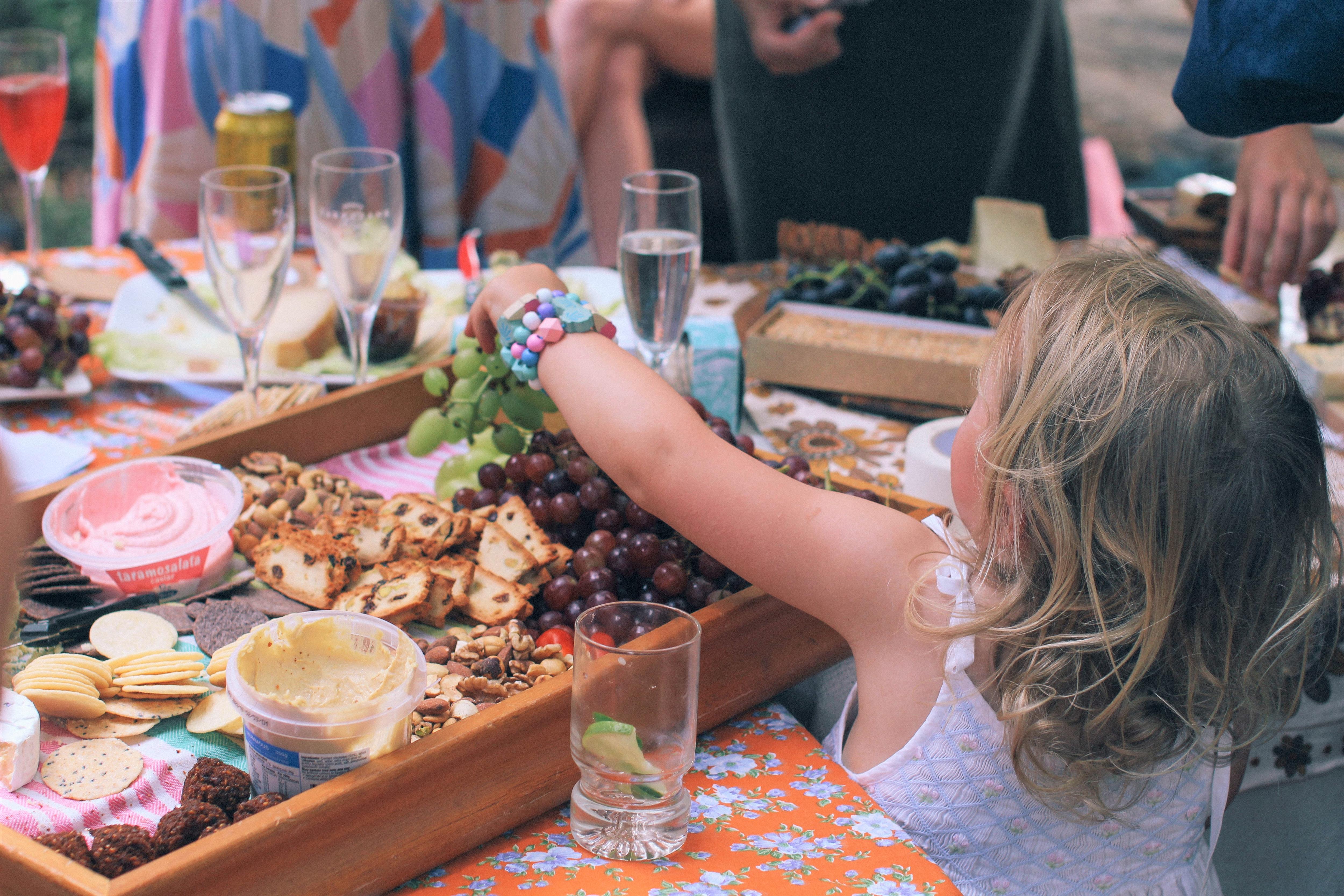 A young child seen from the back reaches up to a table filled with dips and crackers and champagne glasses