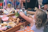 A young child seen from the back reaches up to a table filled with dips and crackers and champagne glasses