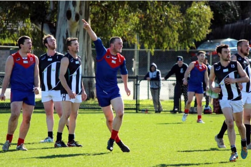 A Lilydale Demons player calls out to a teammate while on a football field surrounded by players from the opposing side.