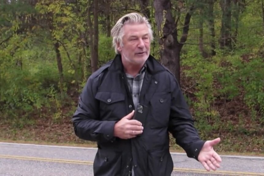 Alec Baldwin speaking on a road with forest behind him.