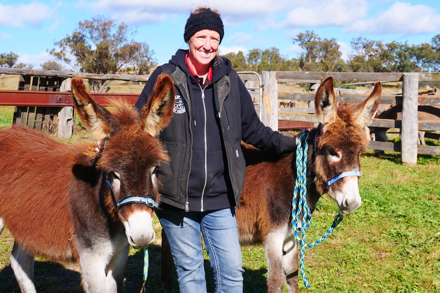 A woman stands between two young donkeys smiling to camera.