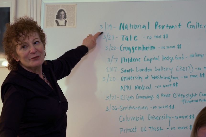 A red-haired woman in her late 60s points at a whiteboard. On that whiteboard is a list of dates and galleries.