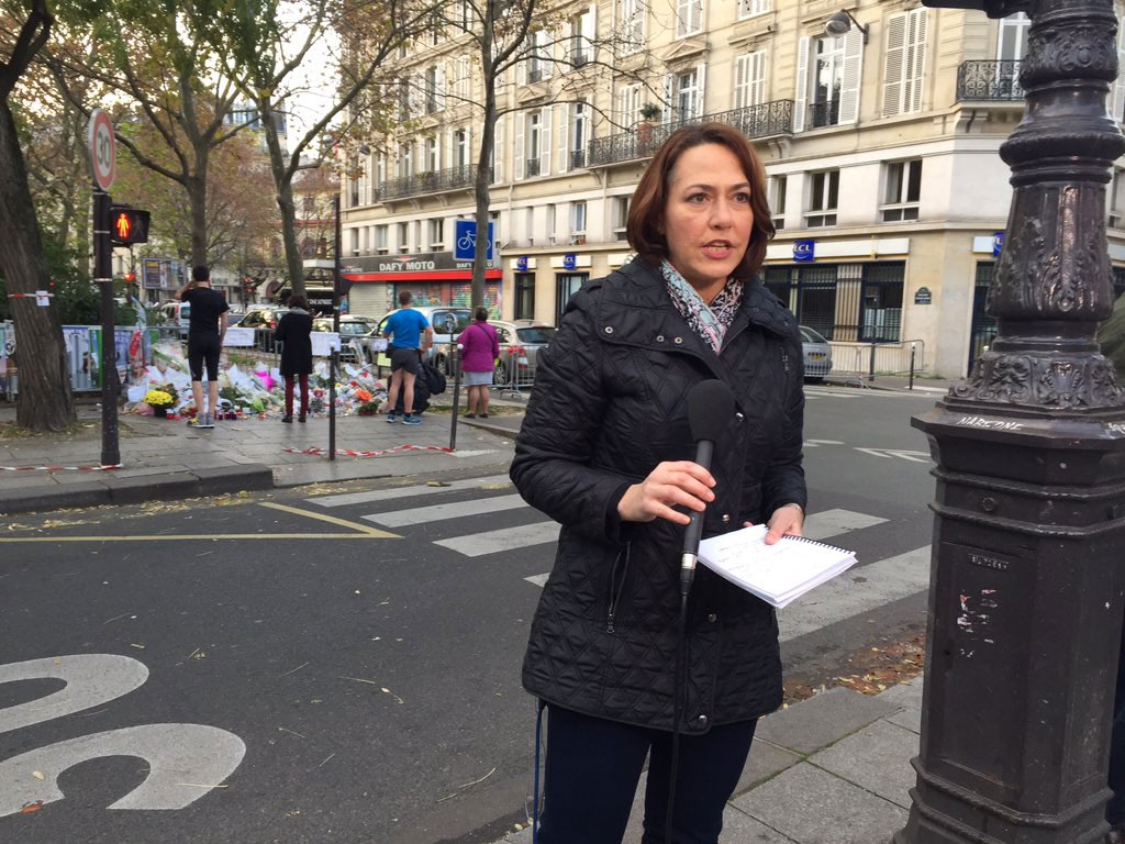 Millar holding microphone and notebook standing in front of floral tributes on street.