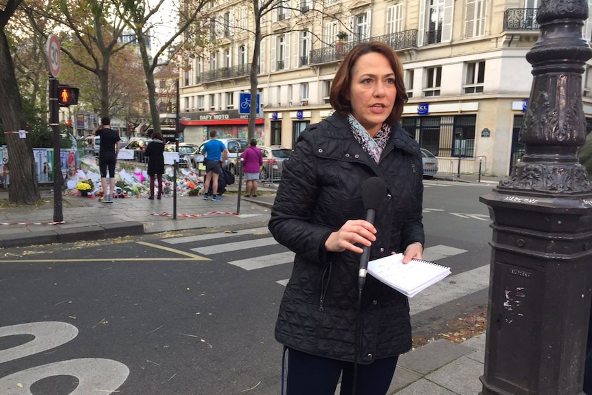 Millar holding microphone and notebook standing in front of floral tributes on street.