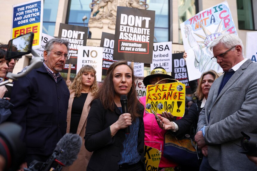 A woman with long dark hair speaks into a microphone in front of a crowd of protesters holding signs.