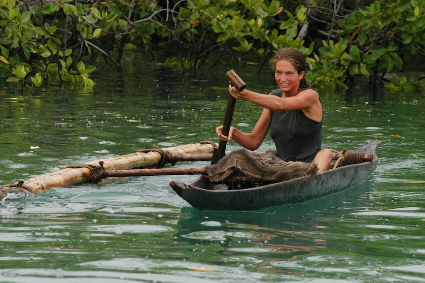 A fit looking woman with brown curly hair kayaks in a river, mangroves growing behind her