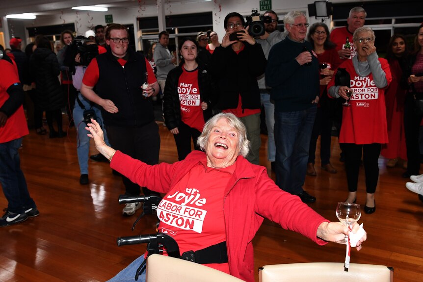An older woman wearing red Labor clothing sits in a walker beaming and holding a wine glass in a crowd of red-shirted people.